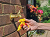 Person holding a yellow garden hose tap adapter connecting to a brass outdoor faucet on a brick wall with flowers in the background.