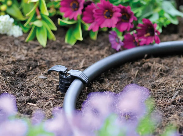 Black garden hose with spray nozzle on soil with flowers in the background