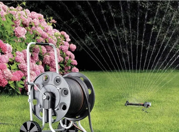 Claber Gemini 8994 Garden hose reel with sprinkler spraying water on grass next to pink flowers