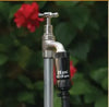 A close-up of a metal garden hose faucet with a black pressure gauge attached, set against a blurred background of green foliage and a red flower.