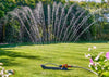 Sprinkler spraying water on a grassy area with flowers and trees in the background