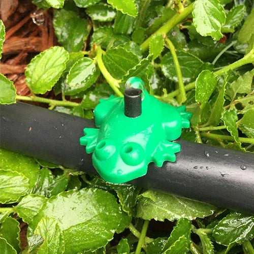 Green frog-shaped garden irrigation dripper on a black pipe surrounded by green leaves