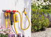 Garden hose with spray nozzle attached to a wall next to a garden scene with flowers and plants.