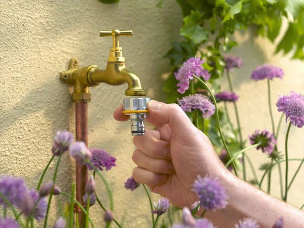 Hand holding a garden hose nozzle near a gold faucet with purple flowers in the background