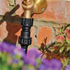 Brass garden faucet with pressure reducer attached to a brick wall with plants in the foreground.