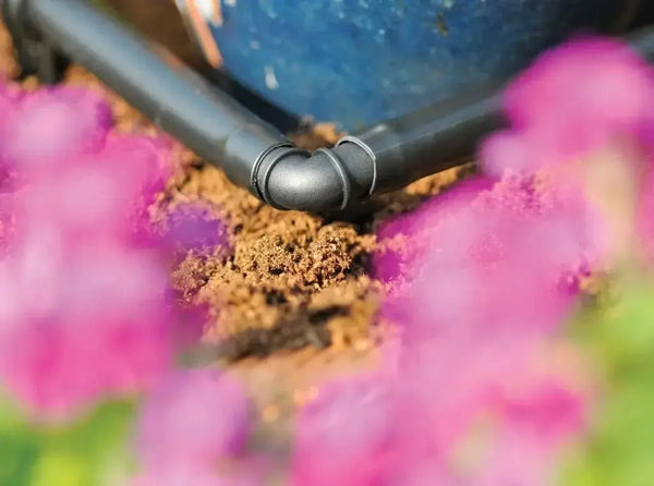 Close-up of a garden irrigation hose elbow connected to a blue container with pink flowers in the foreground.