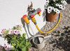 Garden hose with spray nozzle attached to a watering can, with plants and flowers in the background.