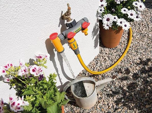 Garden hose with spray nozzle attached to a watering can, with plants and flowers in the background.
