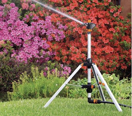 Adjustable tripod garden sprinkler watering green lawn with blooming pink flowers in background
