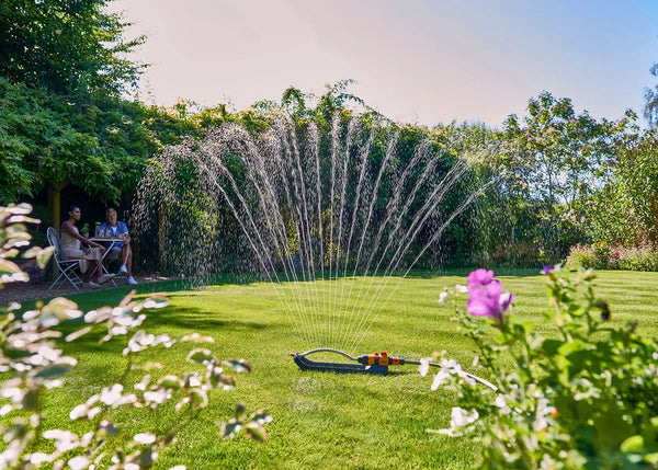Garden with a sprinkler spraying water, people sitting under a tree, and flowers.