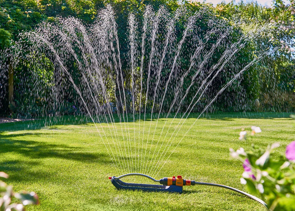 Sprinkler spraying water on a grassy area with flowers and trees in the background