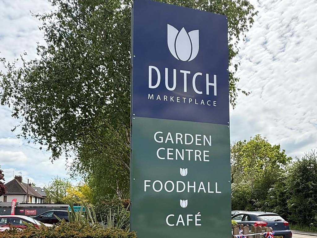 A tall outdoor sign for Dutch Marketplace, with a dark blue top section and a green lower section, featuring white text and tulip logos. Trees, parked cars, and a cloudy sky are visible around the sign.