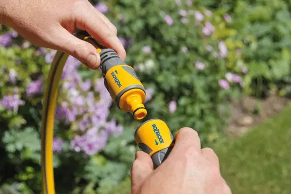 Person holding a yellow and black Hozelock garden hose joiner against a blurred garden background.