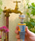 Hand holding a garden hose nozzle attached to a brass faucet with plants in the background